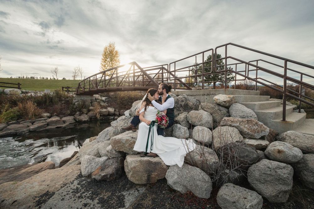 Wedding couple poses on rocky landscape with metal bridge and dramatic sky.