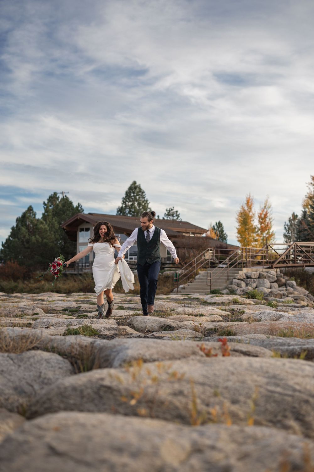 Couple walking hand in hand across rocky terrain with rustic building behind them.