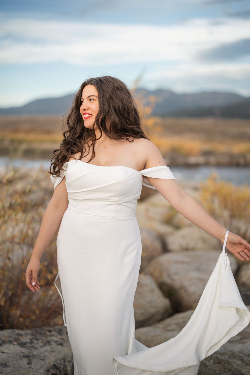 Person in white dress twirling on rocky terrain with mountains and lake in background.