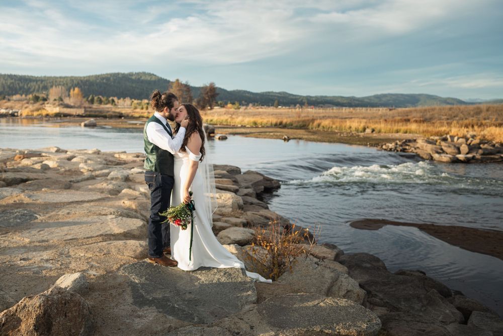 Romantic wedding couple embracing on rocky outcrop beside scenic river with mountains in background.