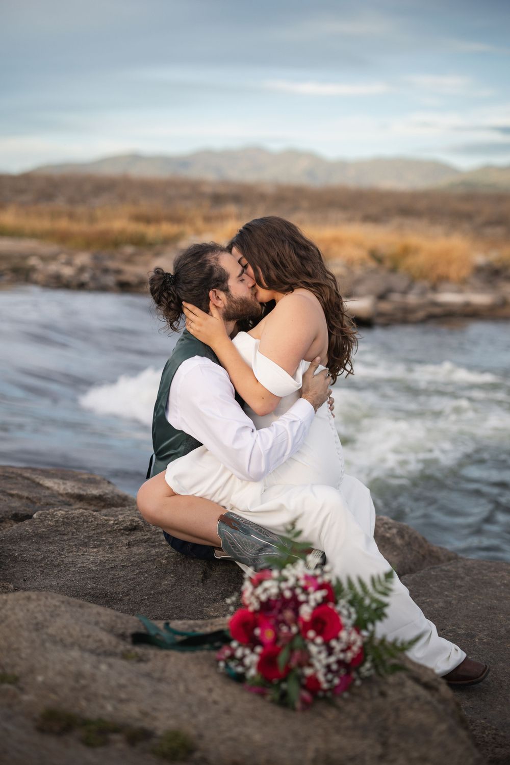 Romantic couple in wedding attire sitting on rocks by flowing river with red bouquet nearby.