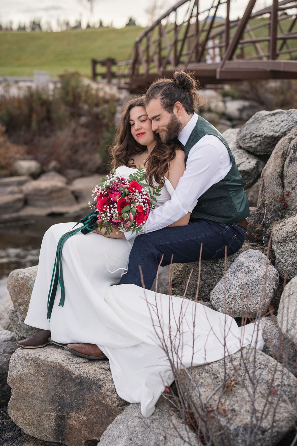 Newlyweds share intimate moment on rocks holding vibrant red bouquet near rustic bridge.