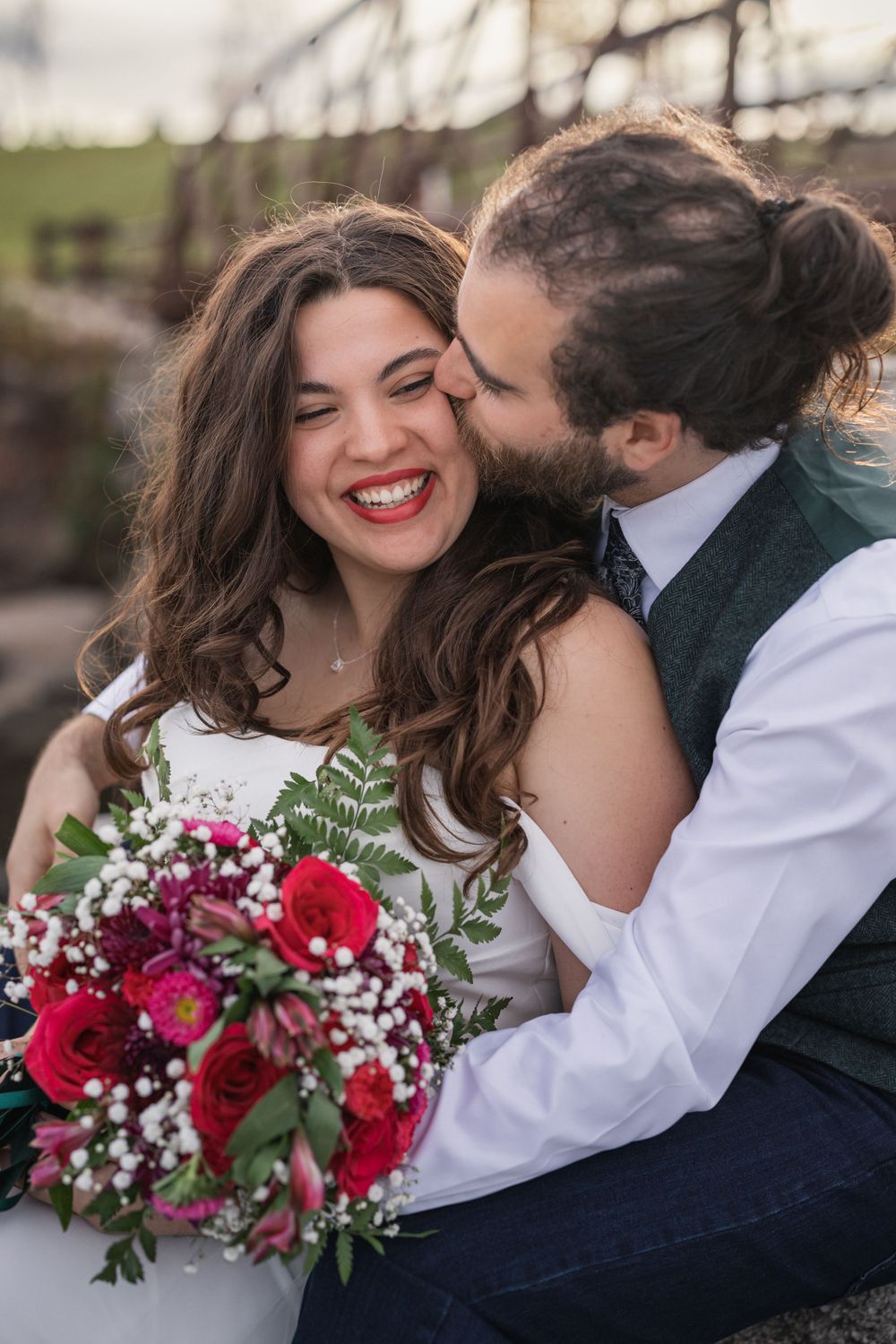 Candid moment of couple laughing with red wedding bouquet by bridge.