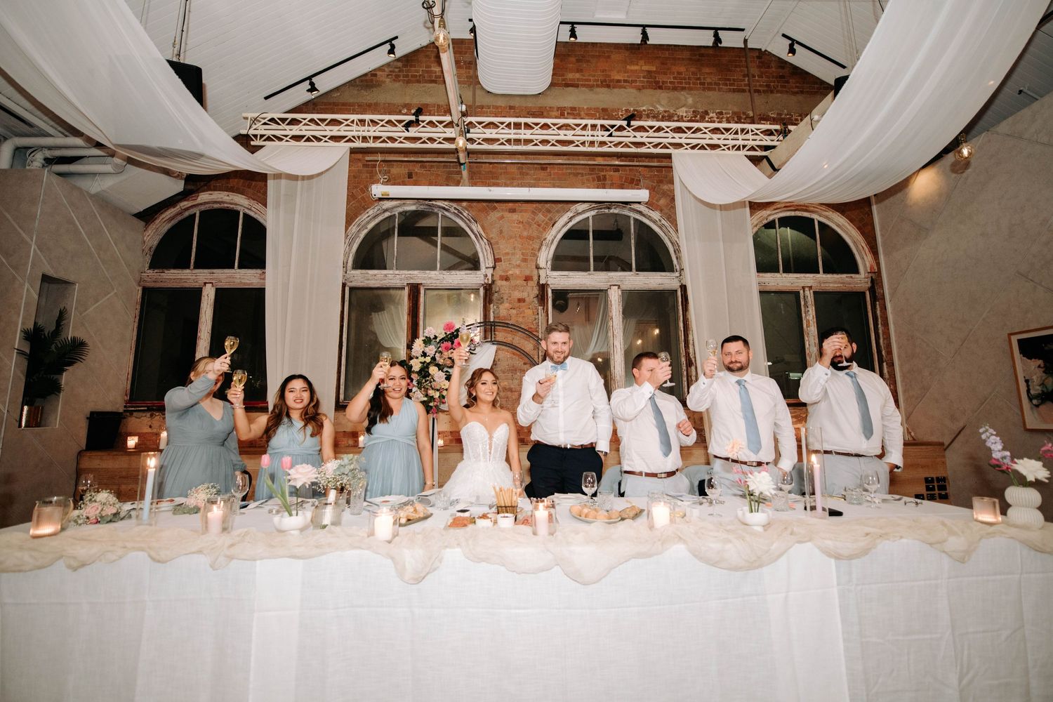 Wedding party members lined up at head table with white draping and exposed brick backdrop.