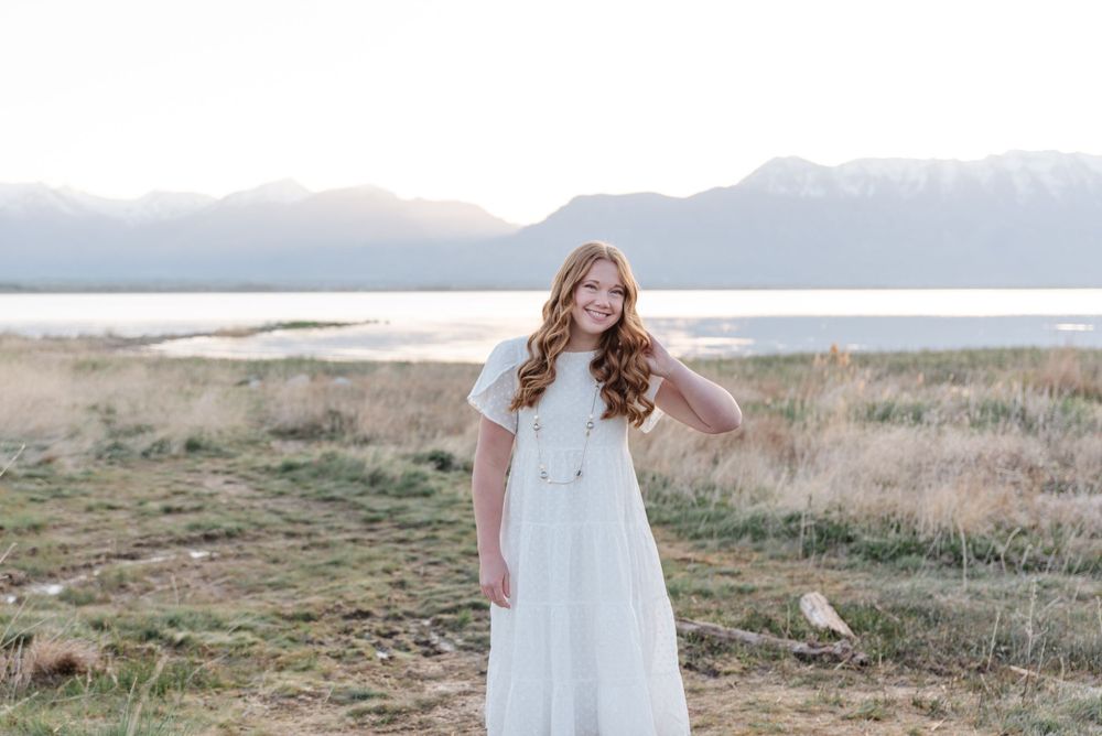 Person in white dress poses in field with mountains and lake in background during sunset.
