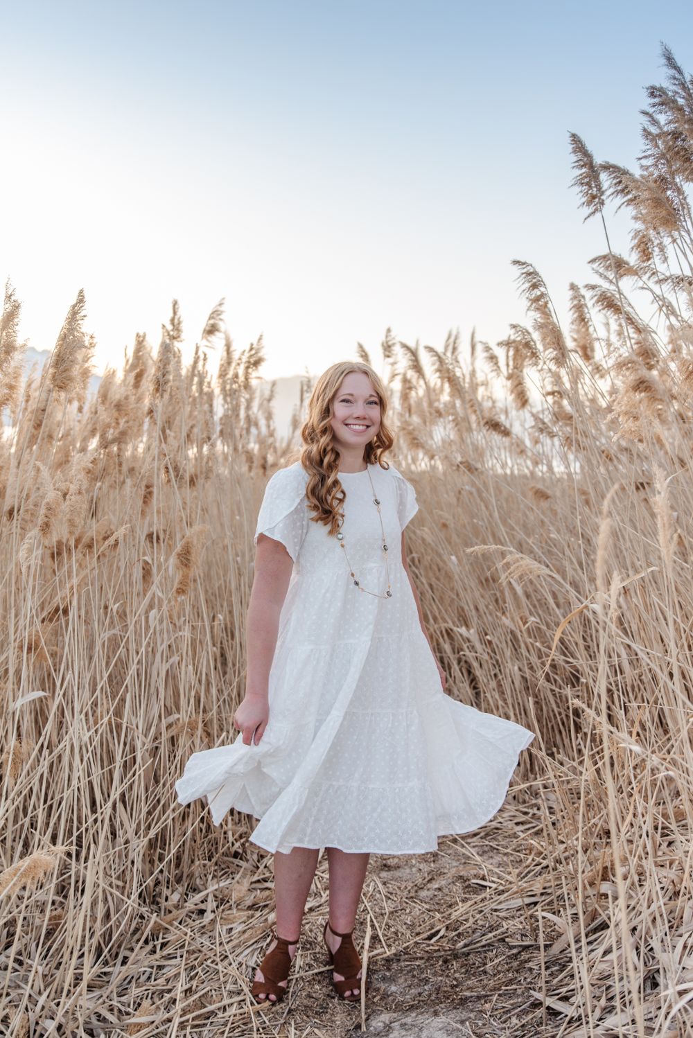 Person in white dress stands among tall golden reeds during sunset.