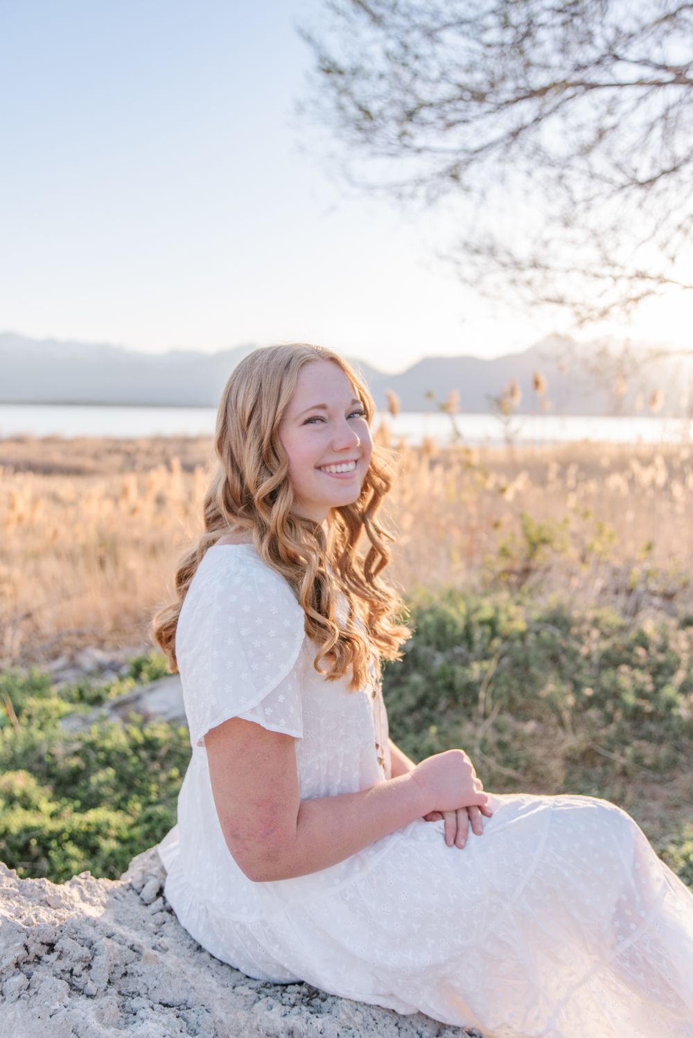 Person in white dress sits on rock during sunset with mountains in background.