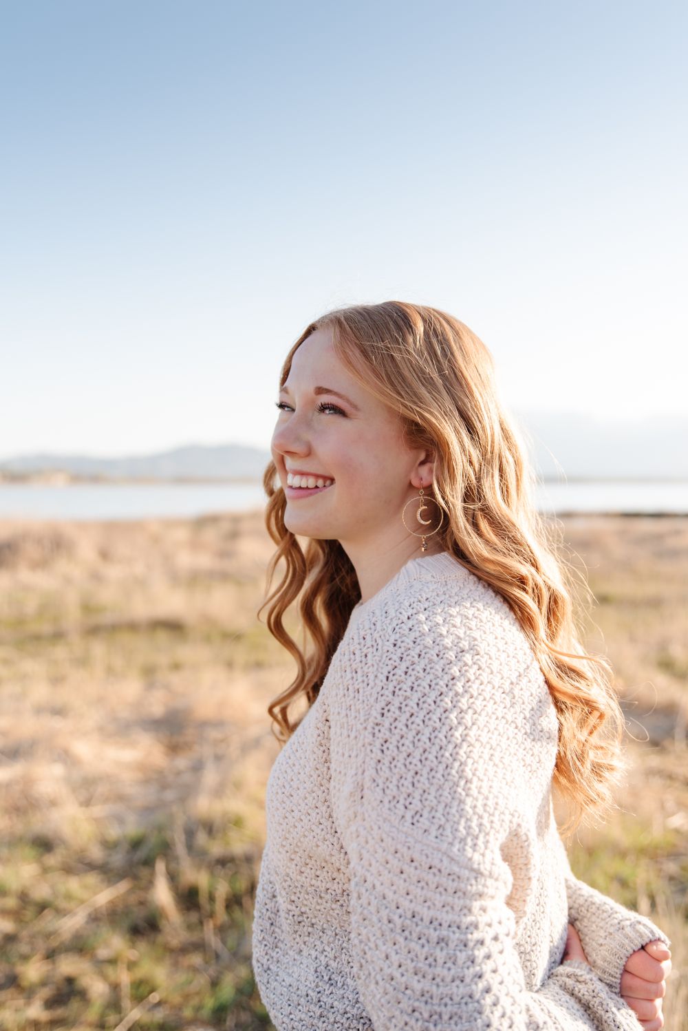 Portrait in a cream sweater with wavy hair looking to the side against a scenic mountain backdrop on a sunny day.