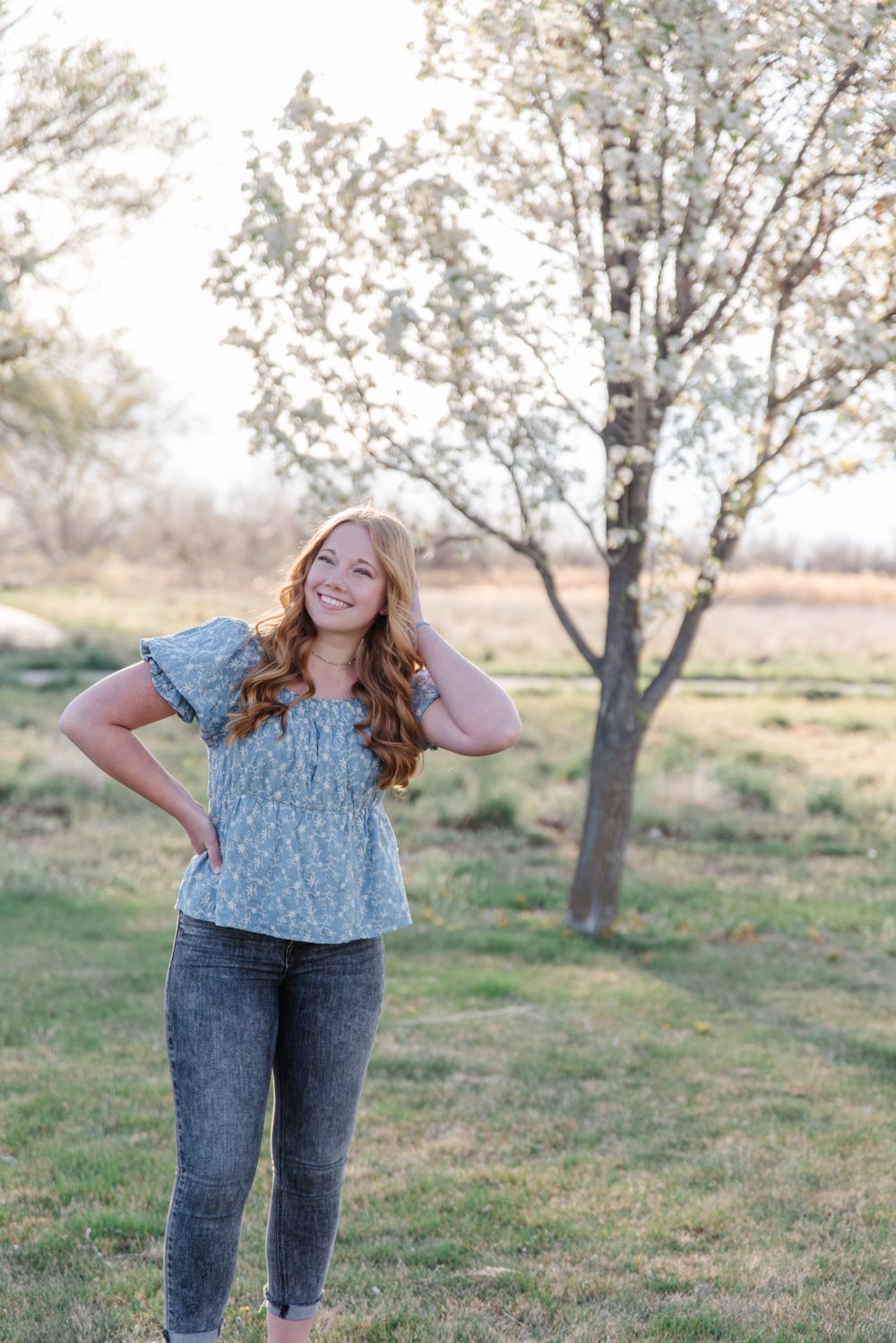 Candid outdoor portrait in a floral top and jeans standing next to a blooming tree.