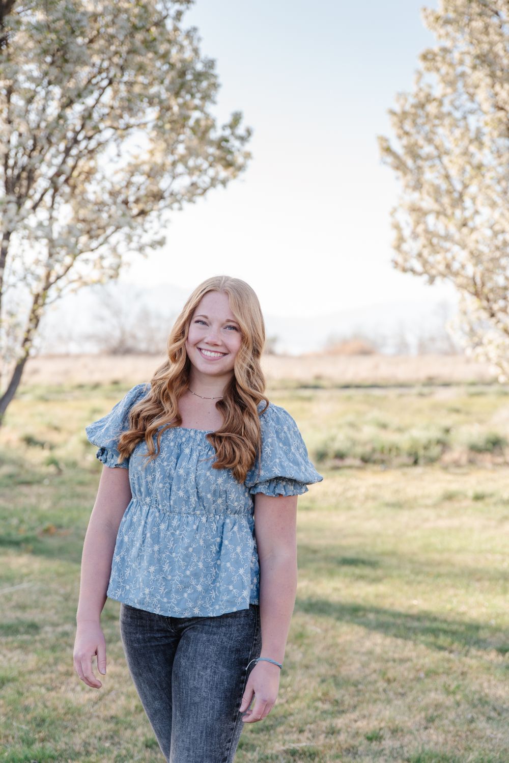 Natural light portrait in a blue floral top and jeans with trees framing the background.