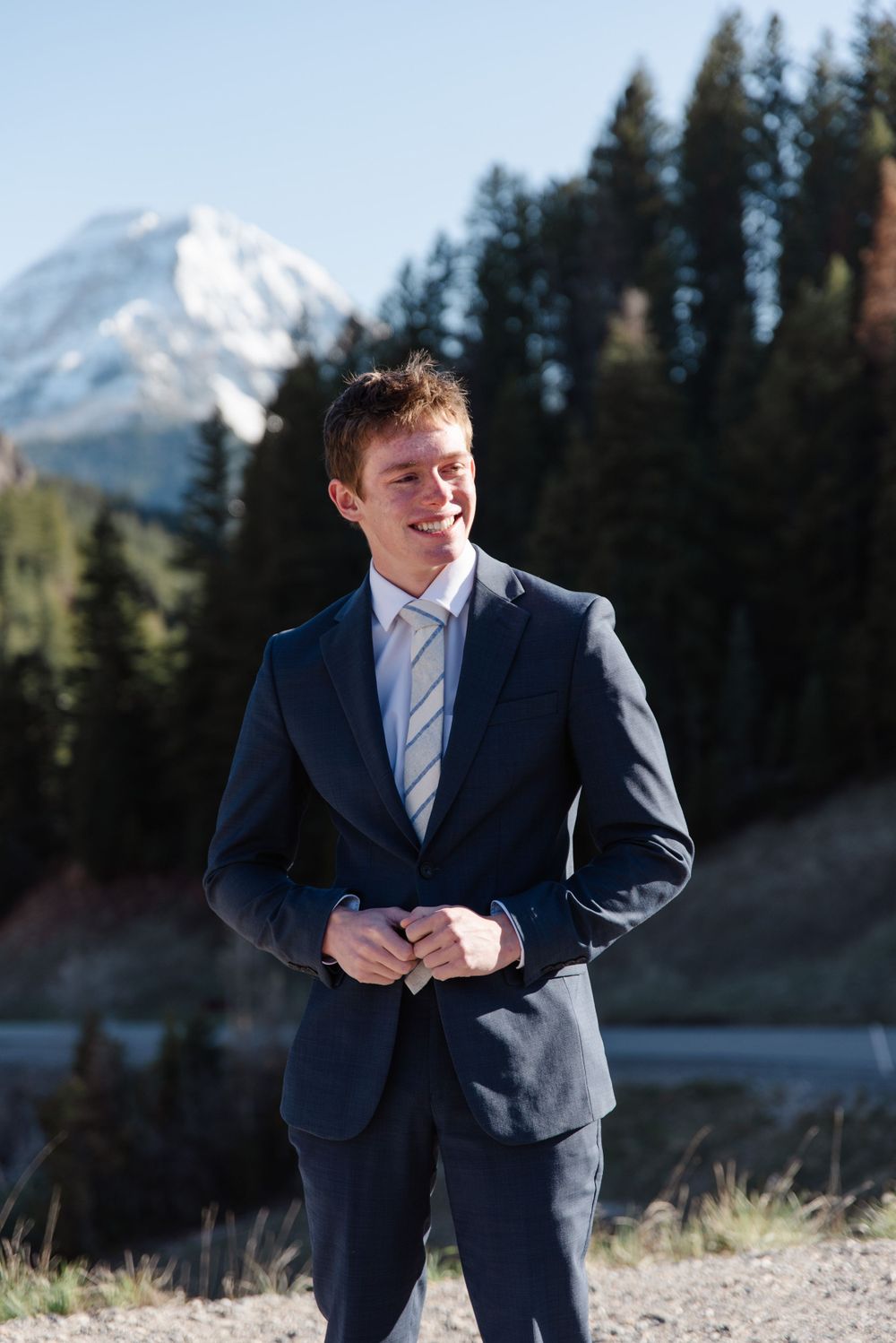 Person in navy suit poses with snowy mountain peak and evergreen forest in background.