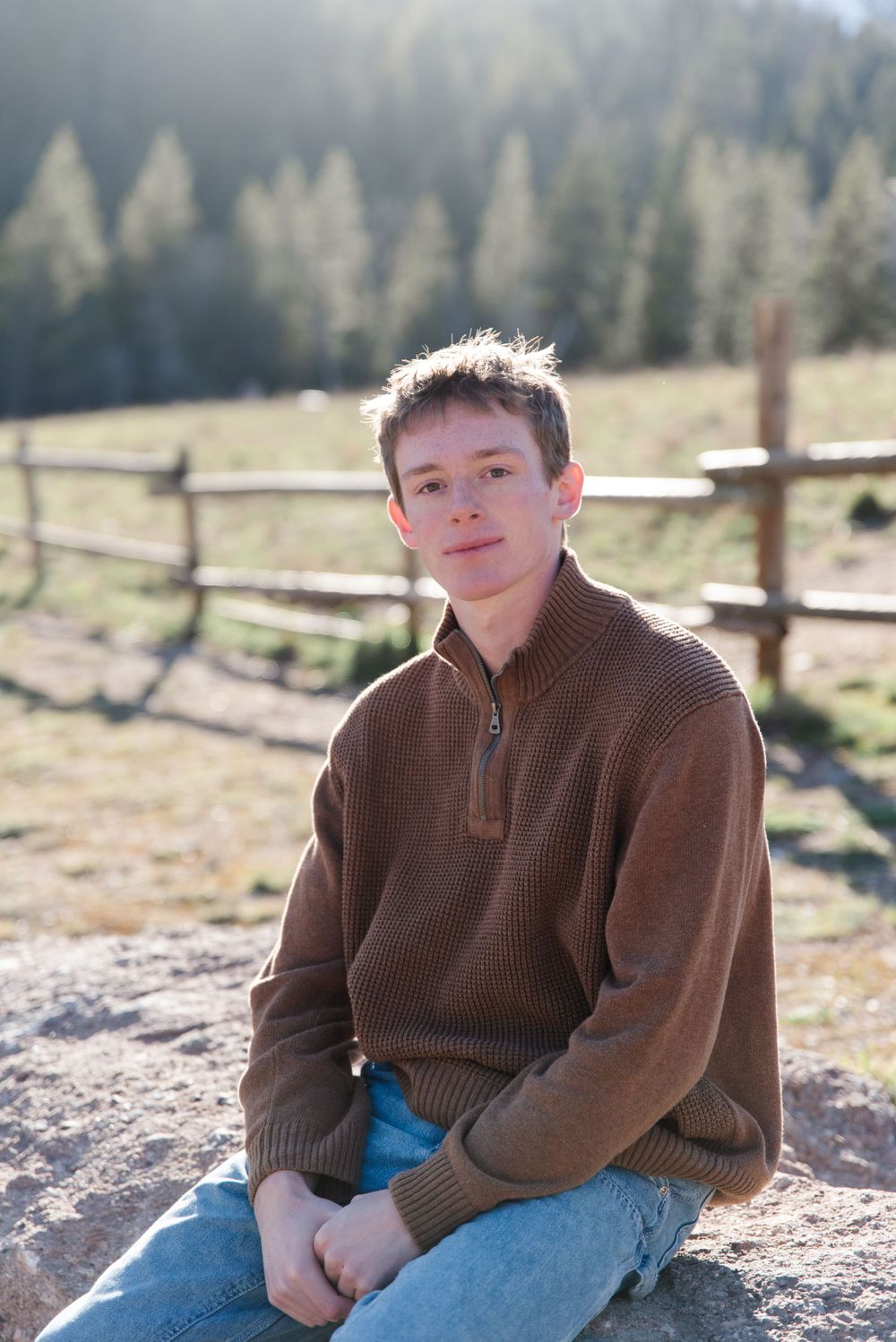 Casual portrait in brown sweater sitting on rock with mountain landscape and wooden fence behind.