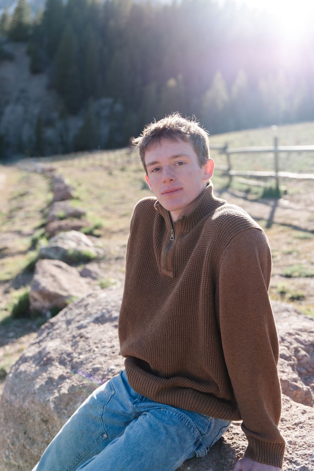 Outdoor portrait in brown knit sweater against sunlit mountain scenery with wooden fence.