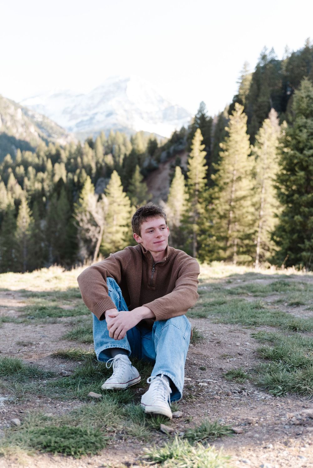 Person in brown jacket sits on ground with mountain landscape and pine trees in background.