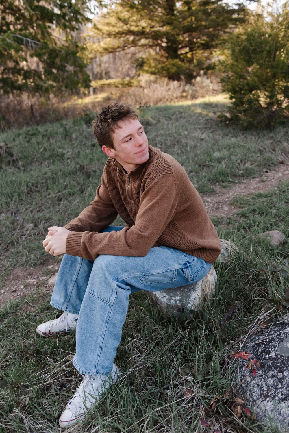 Person in brown sweater and jeans sits on rock in outdoor nature setting.