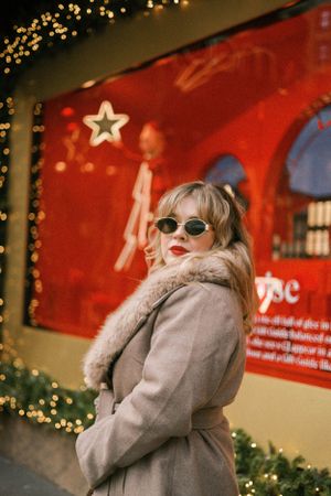 Person in beige coat and sunglasses poses against red wall with holiday lights.