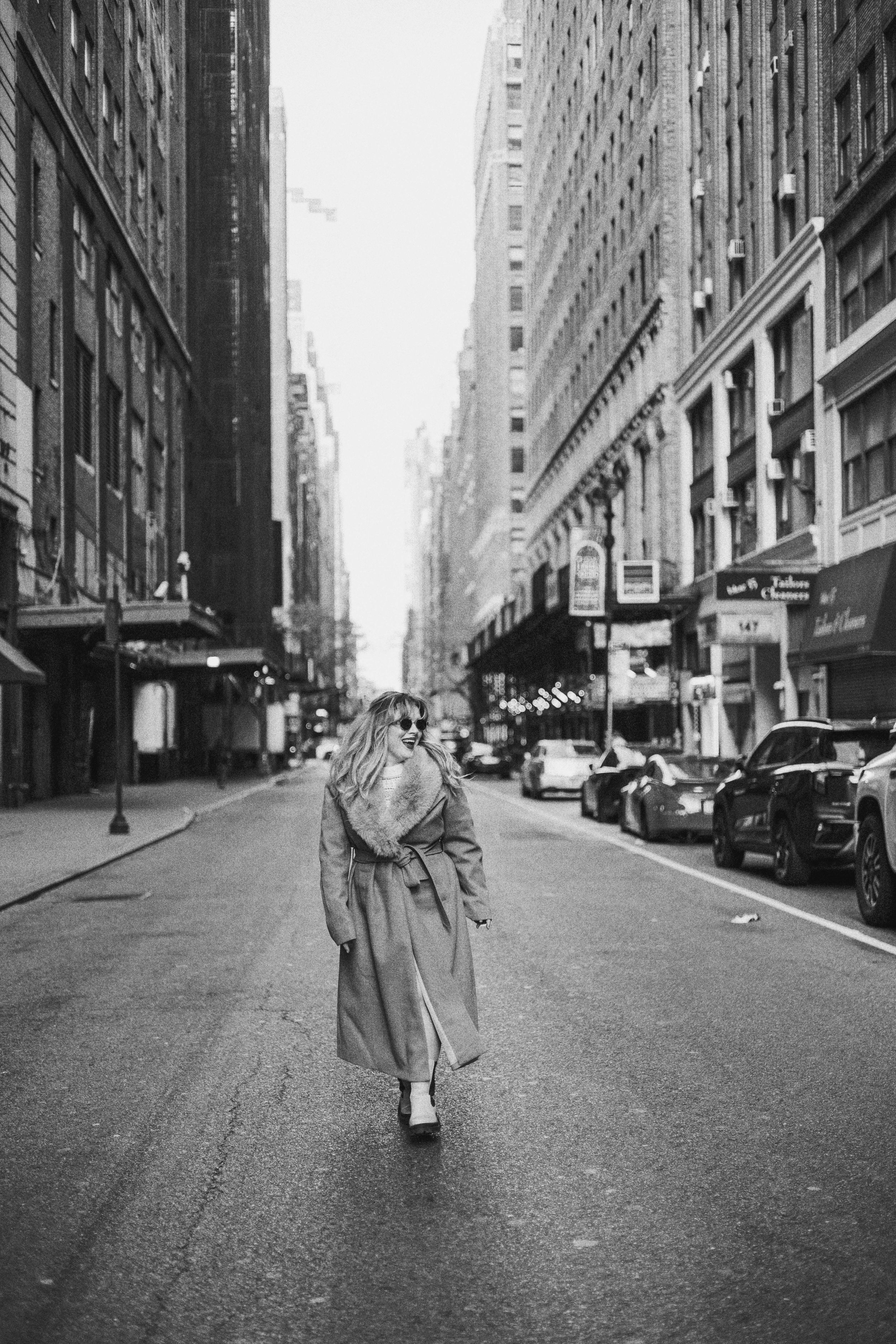 Person walking alone on empty city street wearing long coat in black and white.