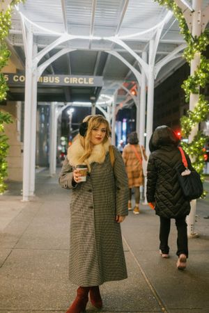 Person holding coffee cup under Columbus Circle archway with holiday decorations.