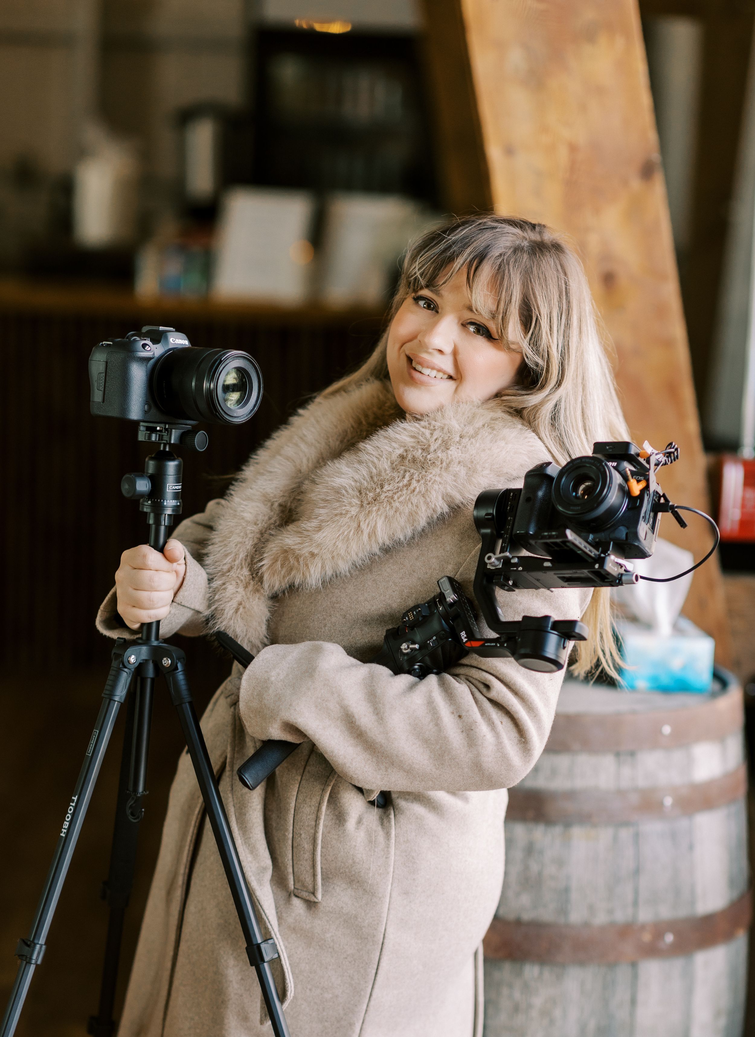 Professional photographer in beige coat with fur collar holding camera equipment and tripod next to wooden barrel.