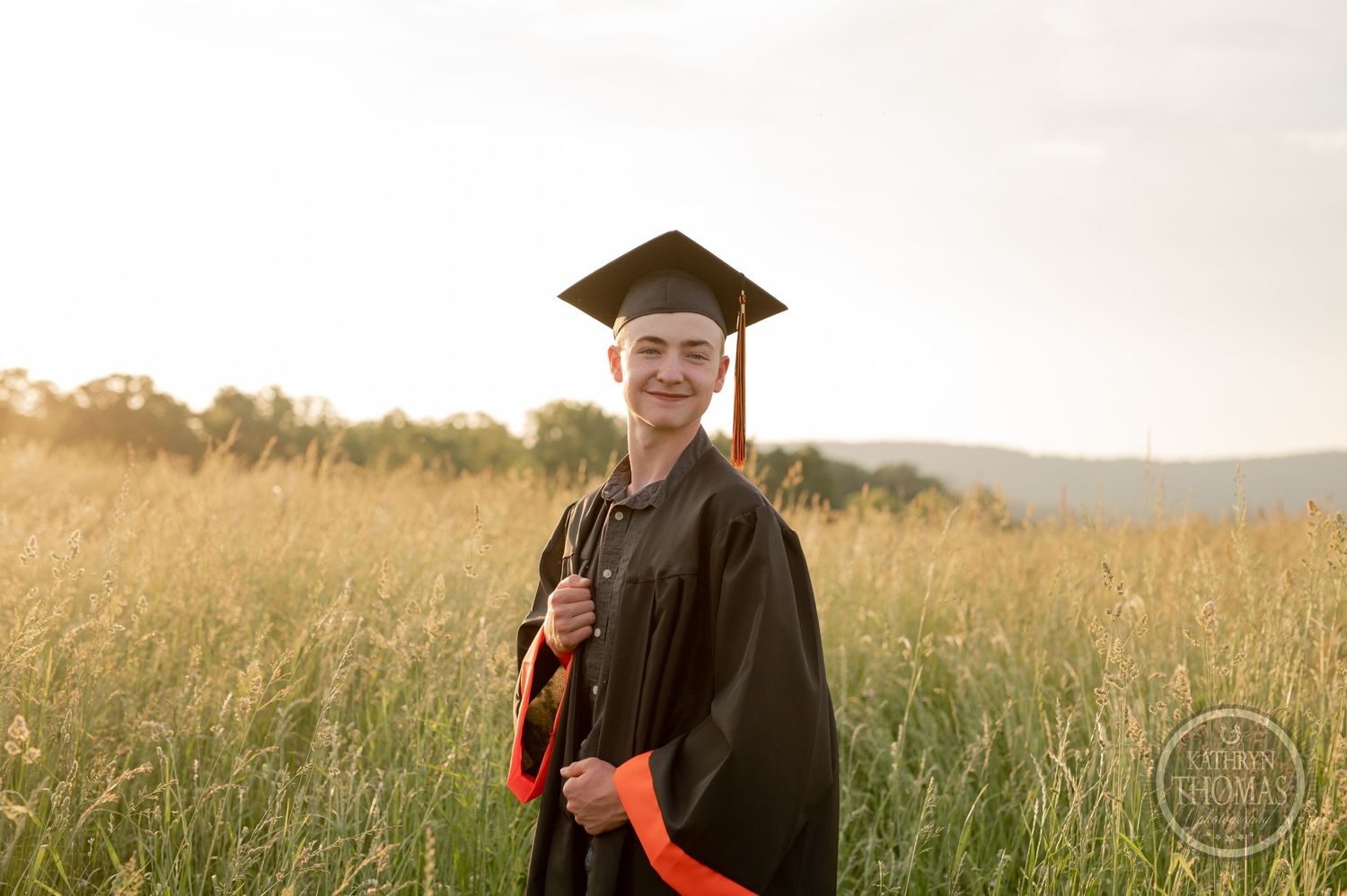 Graduate in cap and gown poses in golden field at sunset with mountains in background.