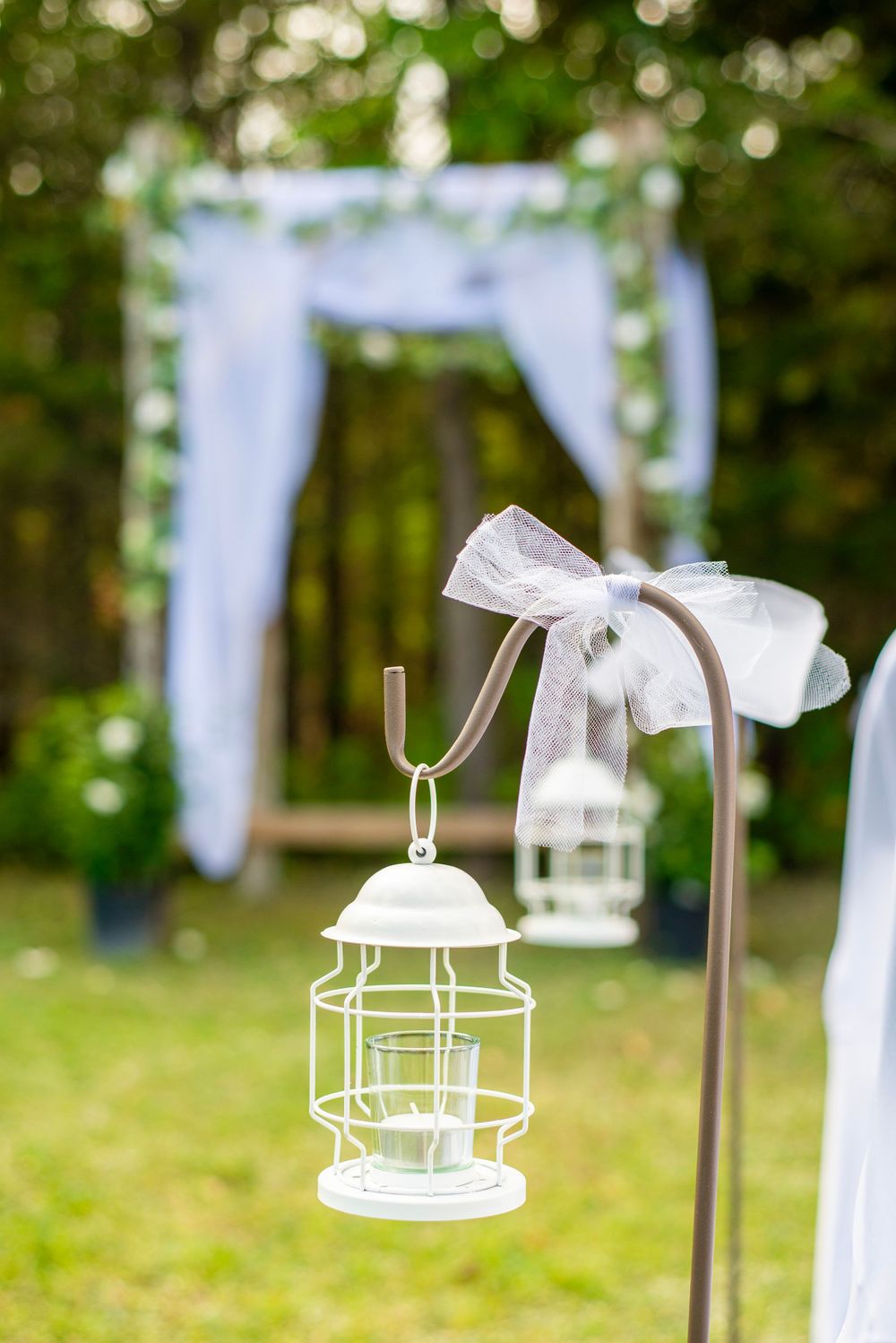 Decorative white lanterns with bows hang along an outdoor wedding aisle with a white arch in the background.