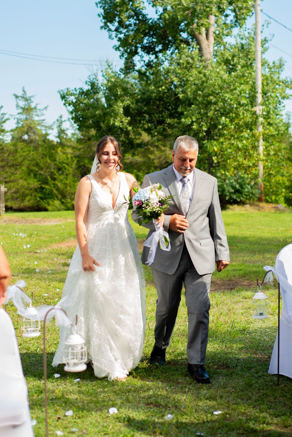 A bride in a beautiful lace gown walks down an outdoor aisle accompanied by someone in a grey suit.