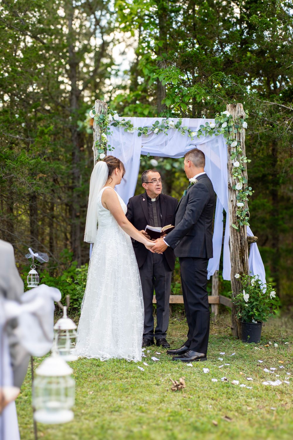 Outdoor wedding ceremony under a white fabric arch decorated with greenery and lanterns in a wooded setting.
