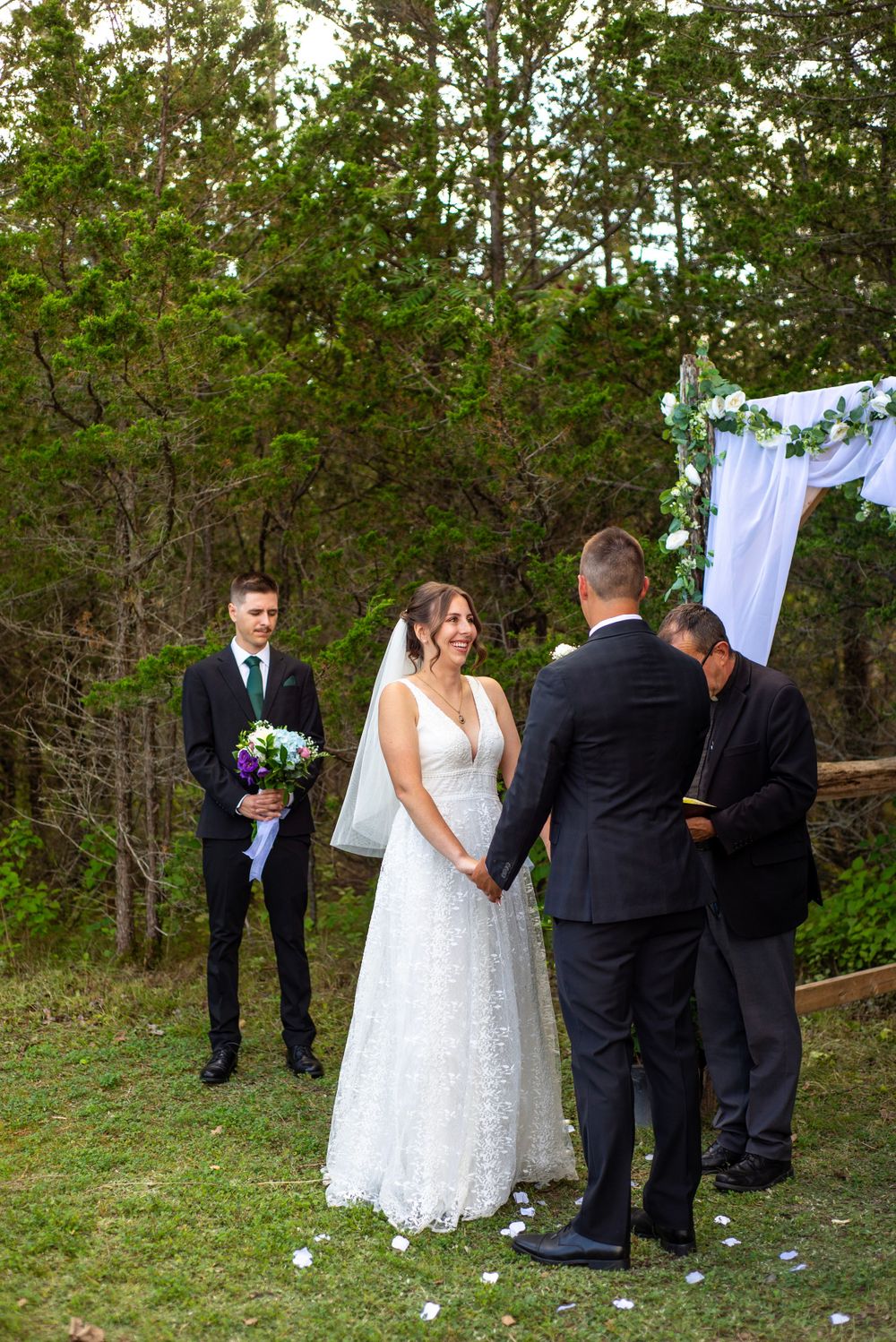 An outdoor wedding ceremony setup with white fabric arch and floral decorations in a wooded setting.
