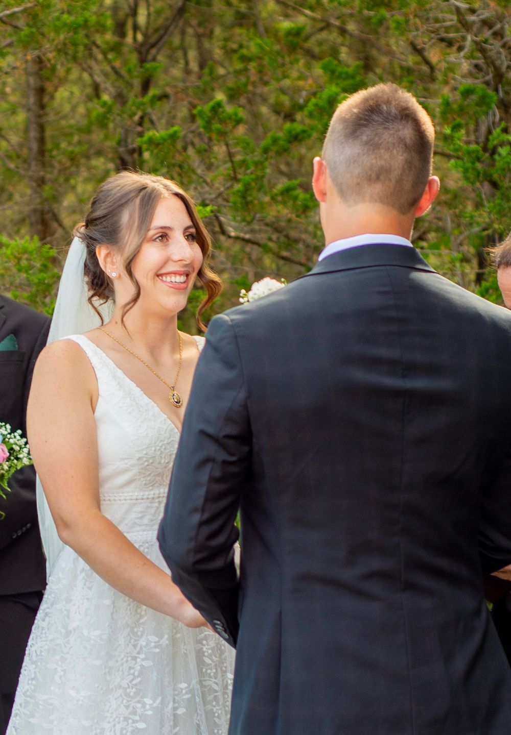Couple exchanging smiles during outdoor wedding ceremony with greenery in background.