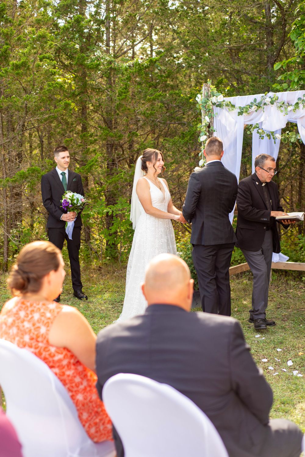 Outdoor wedding ceremony under a white arch with guests seated on covered chairs.
