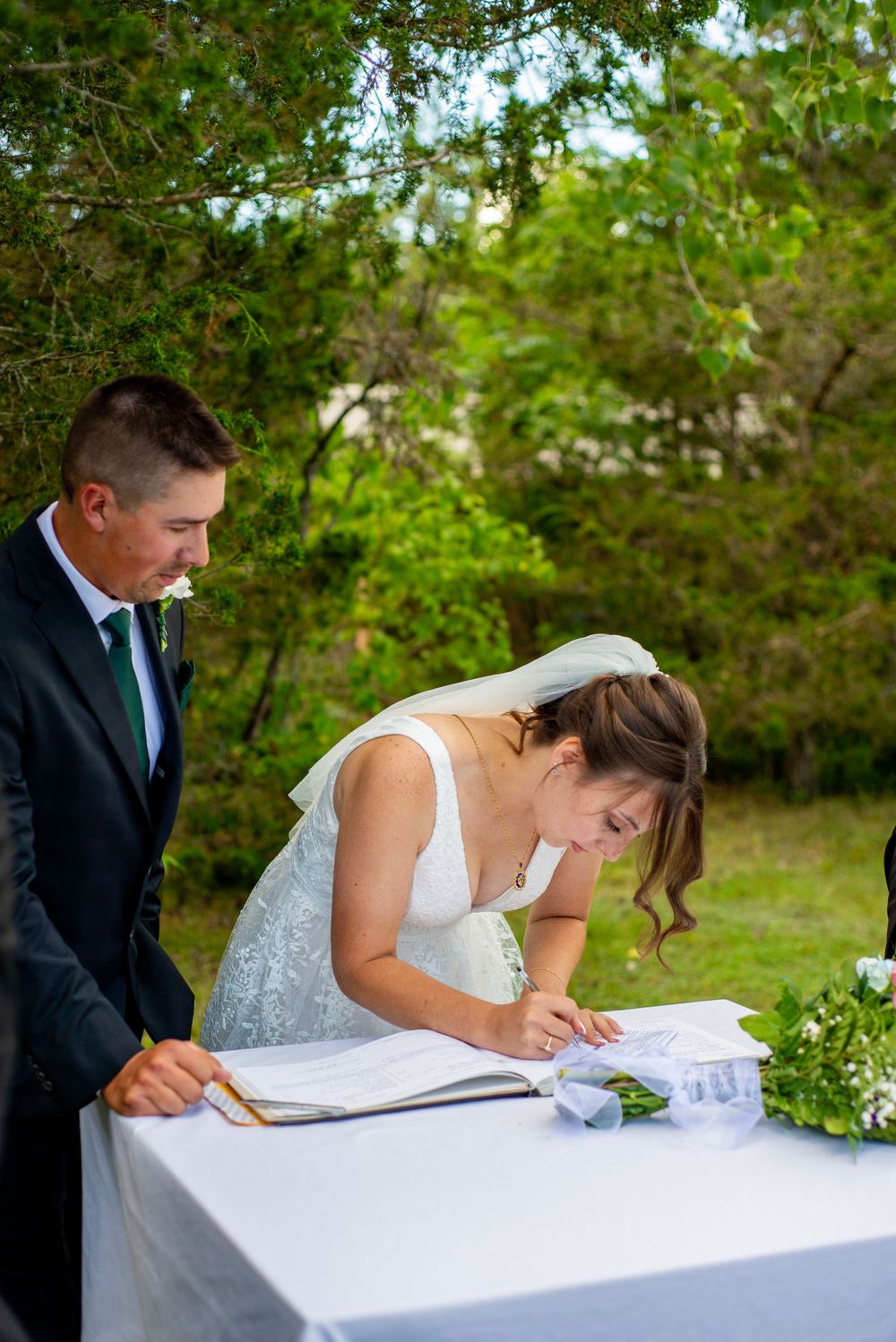 A couple at an outdoor wedding ceremony signing documents at a white-clothed table.