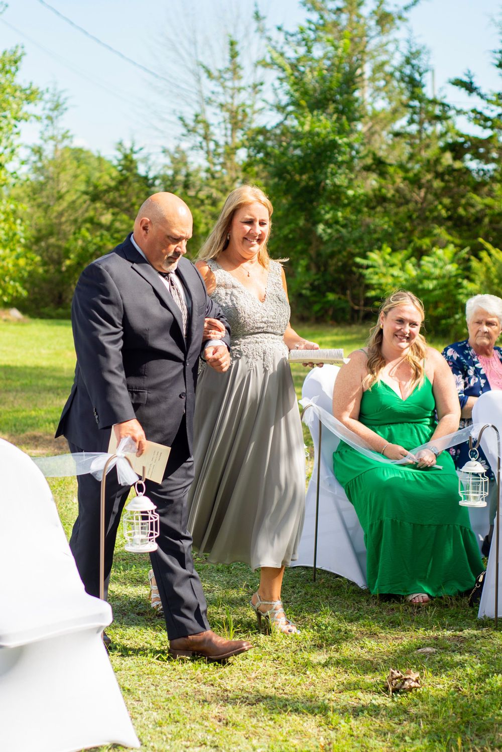 Wedding guests in formal attire gather outdoors on a sunny day with white chairs and green foliage.