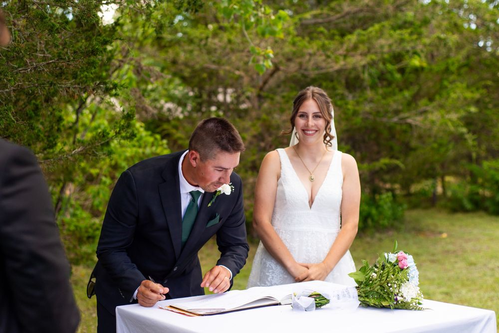 Wedding couple at outdoor ceremony signing marriage license with bridal bouquet on table.