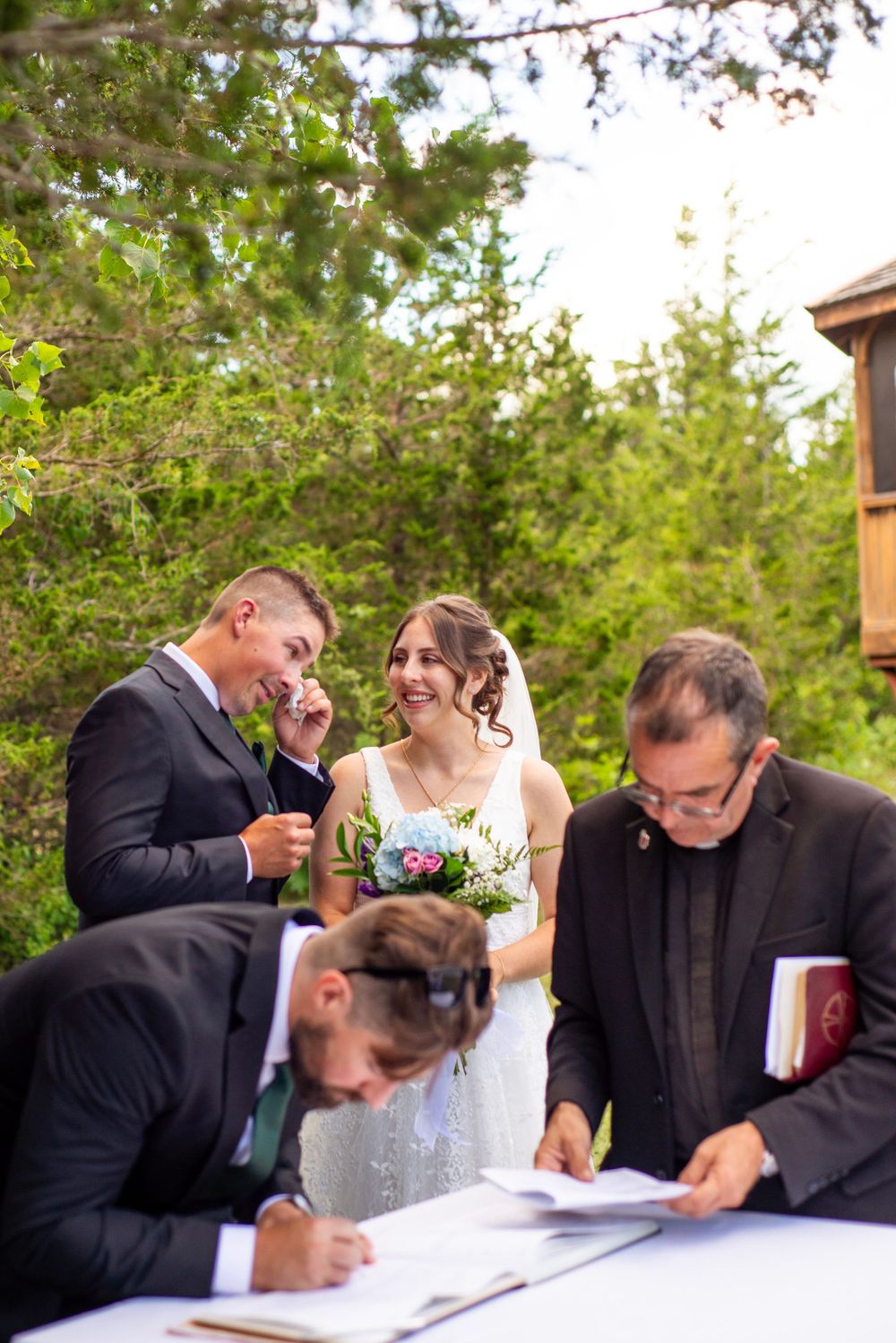 A joyful wedding ceremony moment as documents are being signed at an outdoor venue surrounded by greenery.