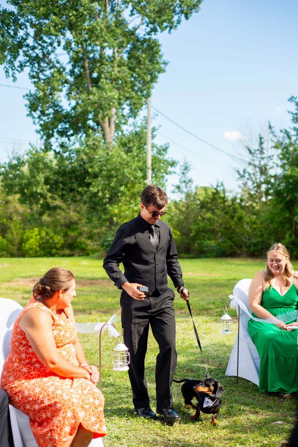 Outdoor wedding scene with attendees and a small dog on lawn with tall trees in background.