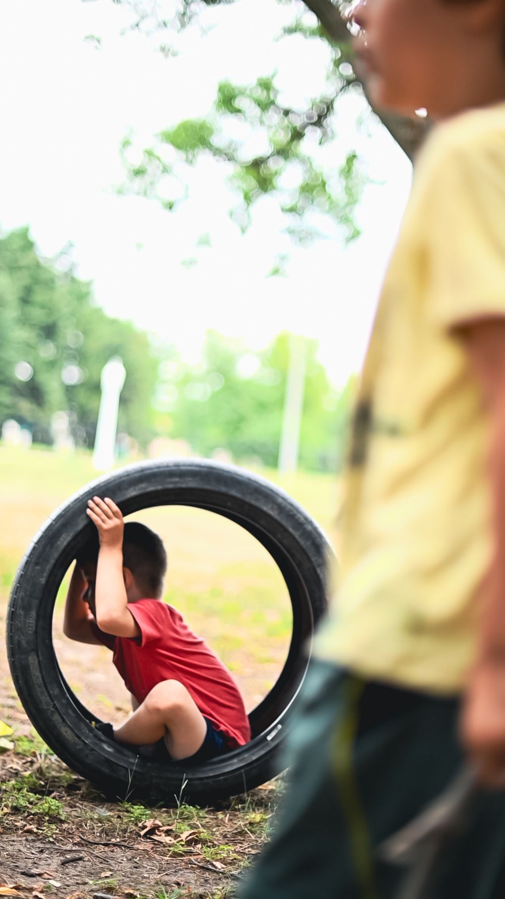 Children playing with large tire outdoors in natural setting with trees in background.