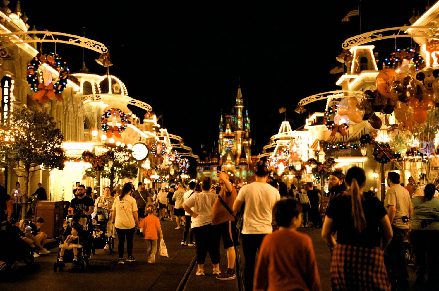 Crowds walk down Main Street USA at Disney World with holiday decorations and castle illuminated at night.