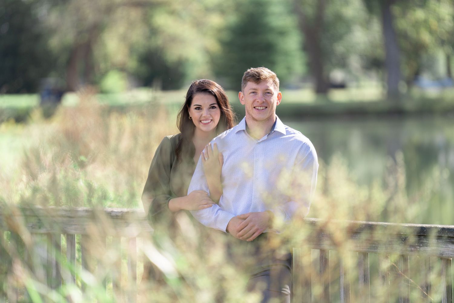 A couple poses together in a romantic outdoor setting with tall grass and a lake in the background.