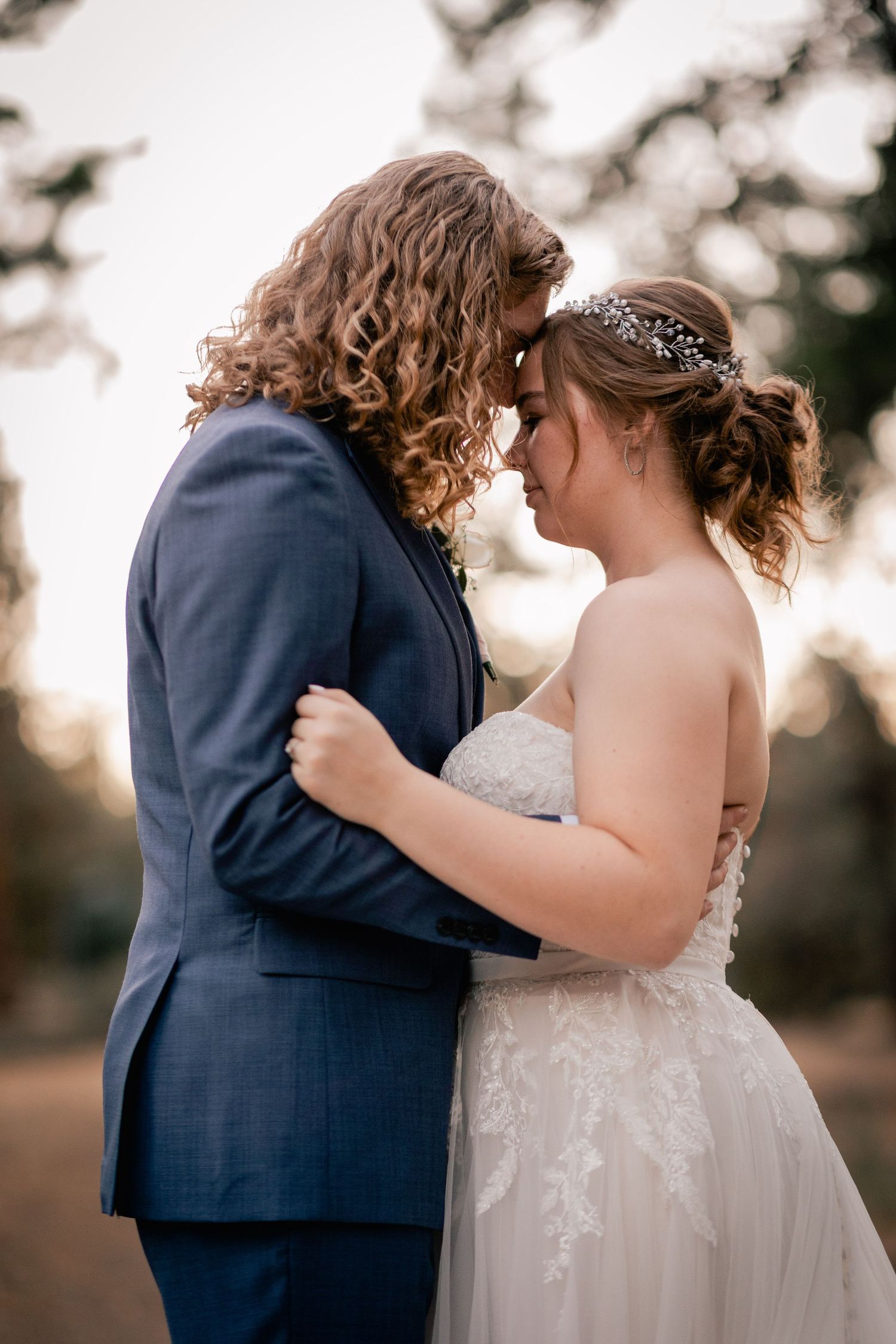 A romantic wedding couple shares an intimate moment at sunset, with the bride in a lace gown and groom in navy suit.