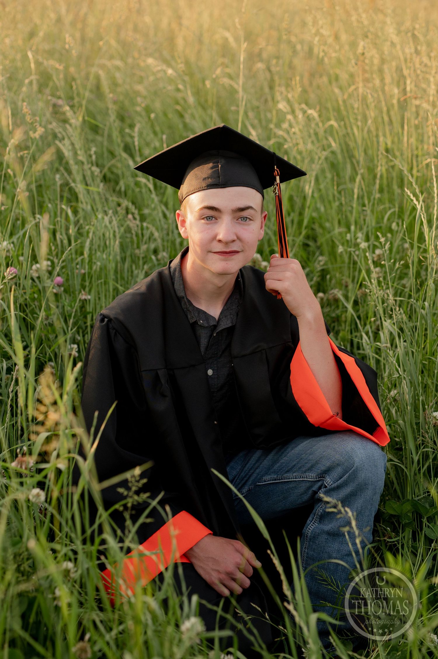 Graduate in cap and gown kneels in tall grass field with orange stole.