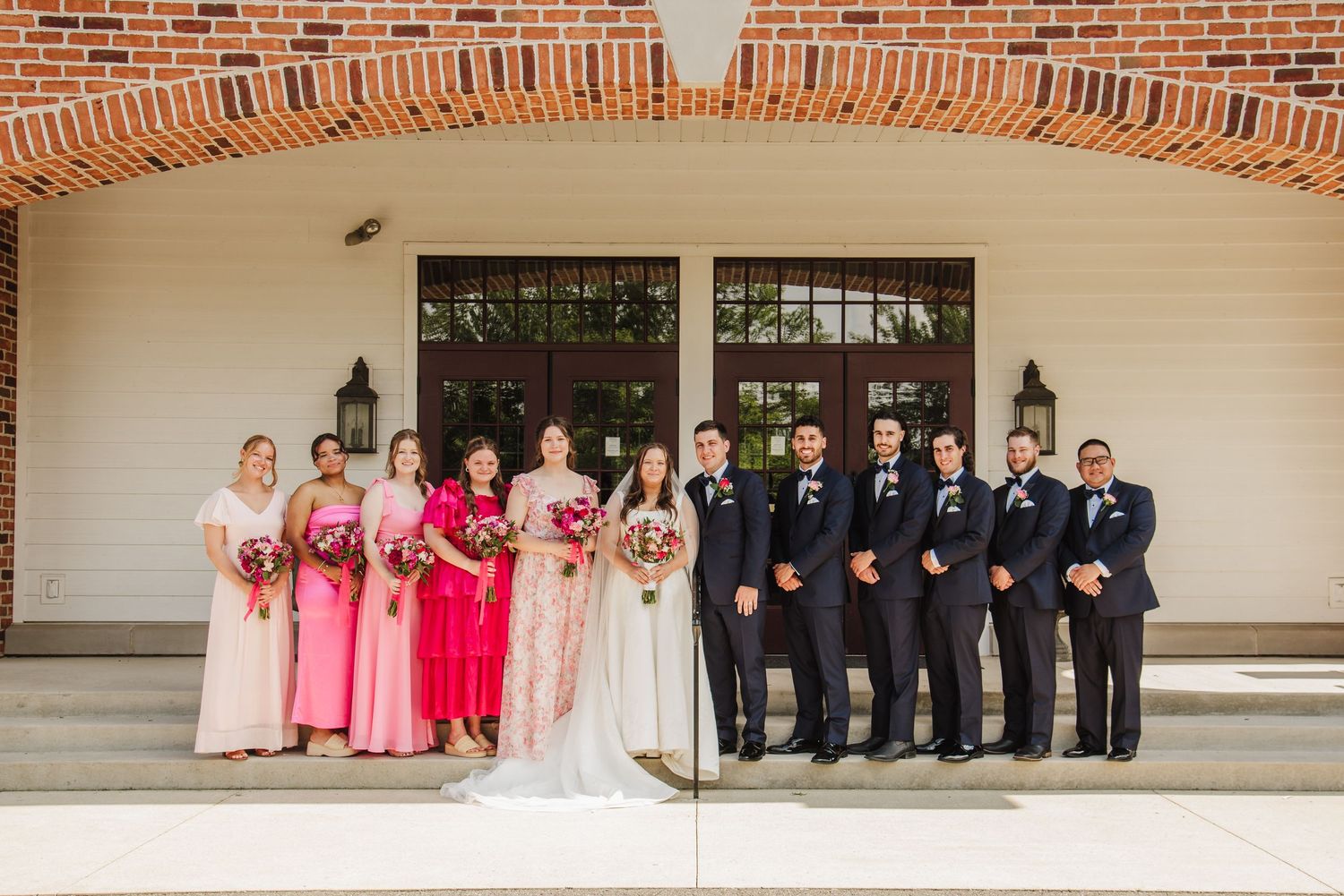 Wedding party lined up against brick wall with bridesmaids in pink dresses and groomsmen in dark suits.