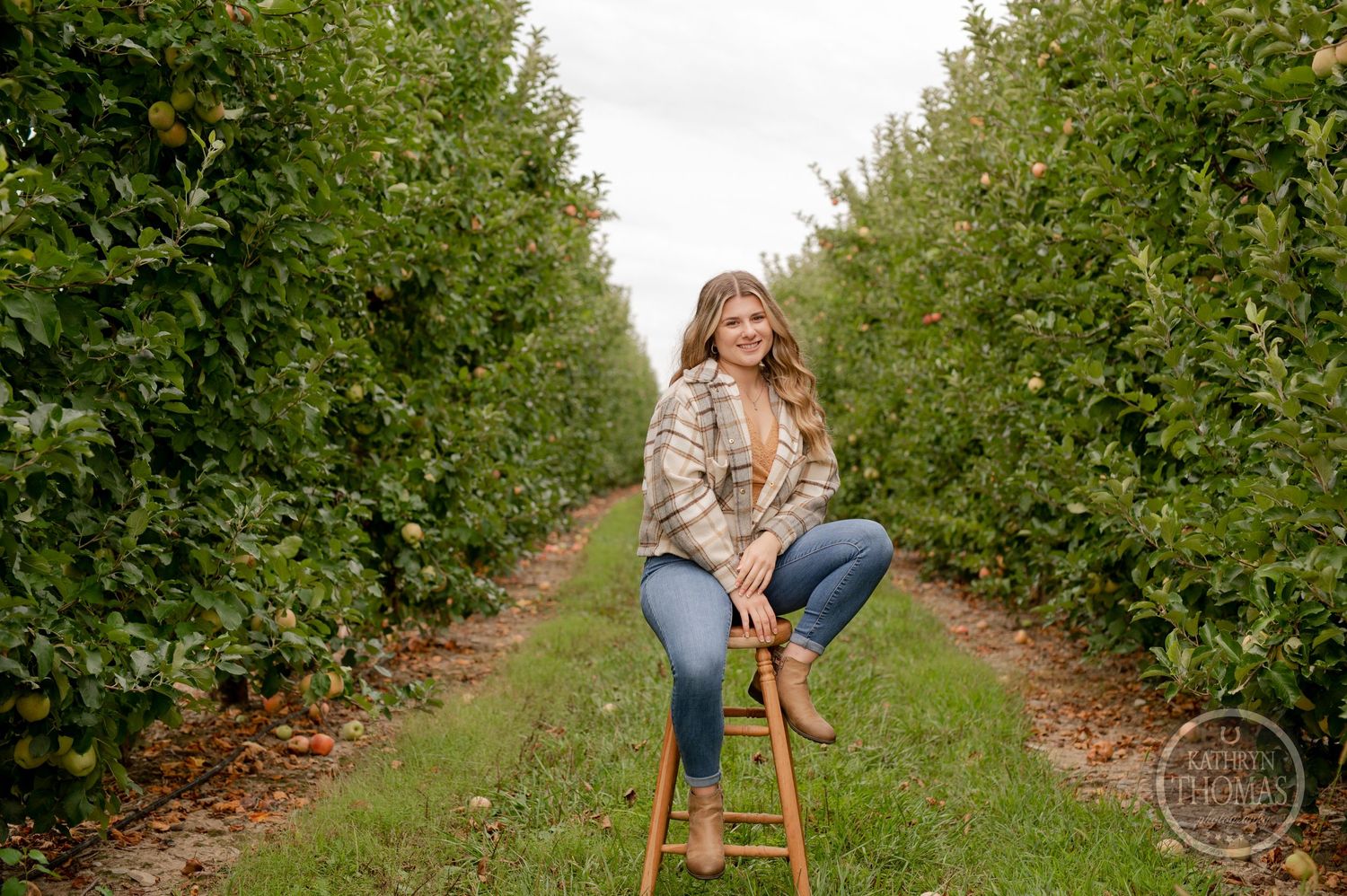 Person posing on wooden stool between rows of apple trees in autumn orchard setting.