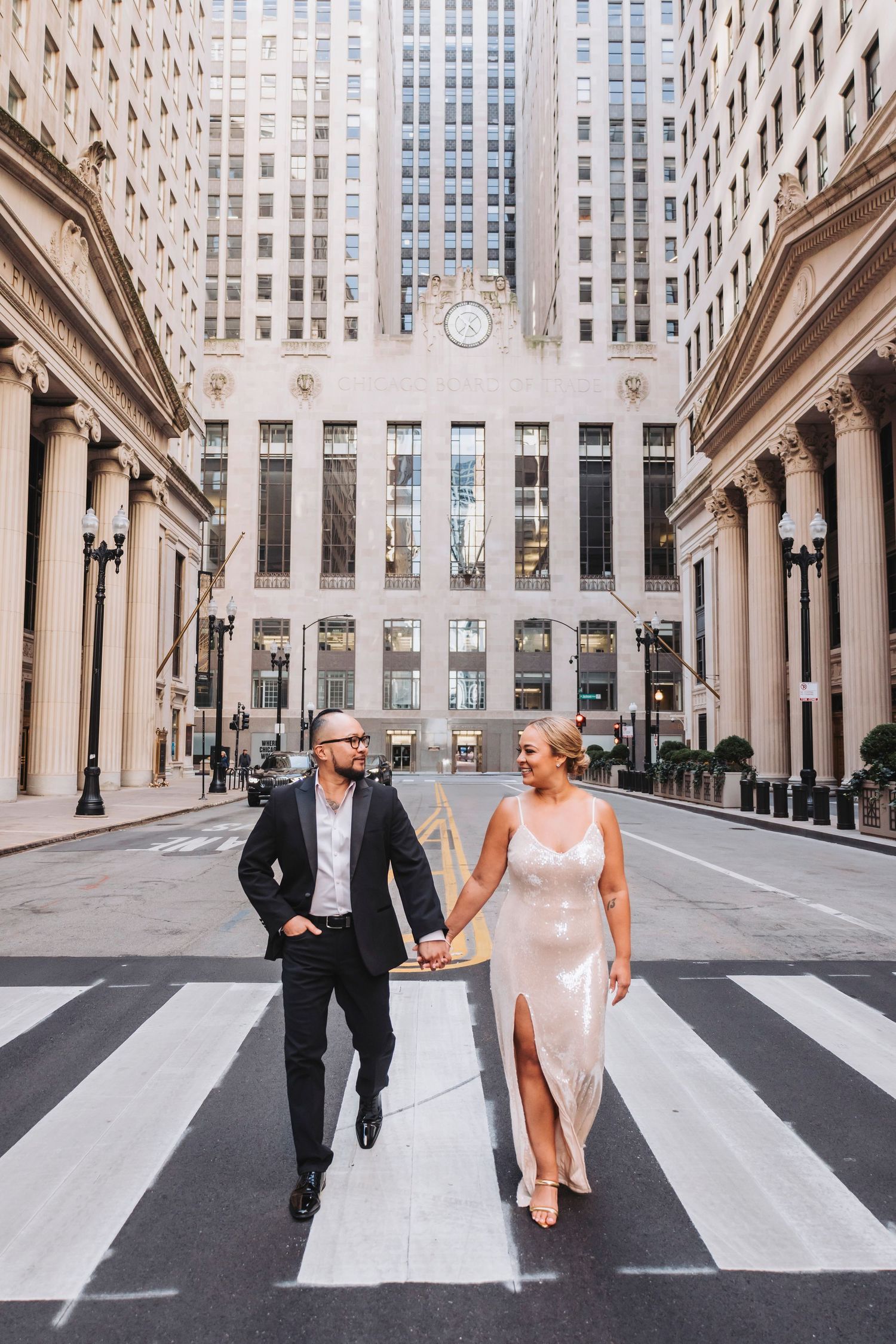 A couple in wedding attire crosses a city street with tall buildings in the background.