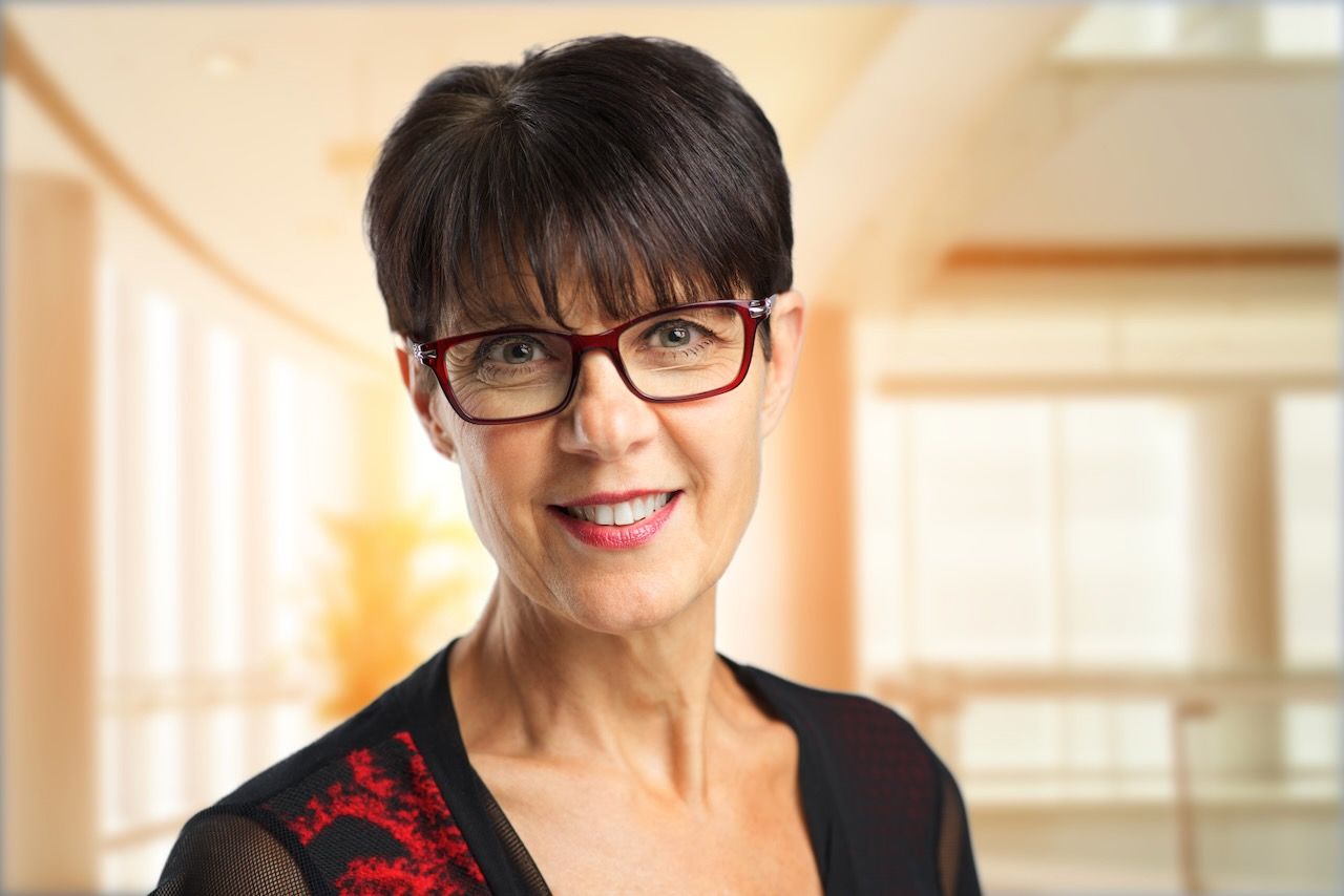 Professional headshot of someone in glasses and a black top with red embroidery detail against a warm blurred background.