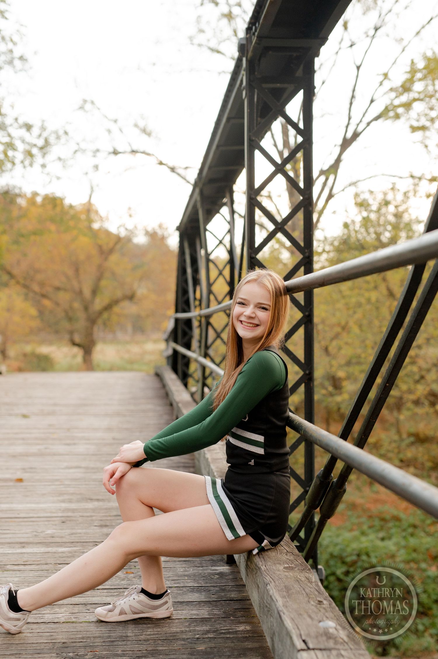 Cheerleader sits casually on bridge railing during autumn photoshoot outdoors.