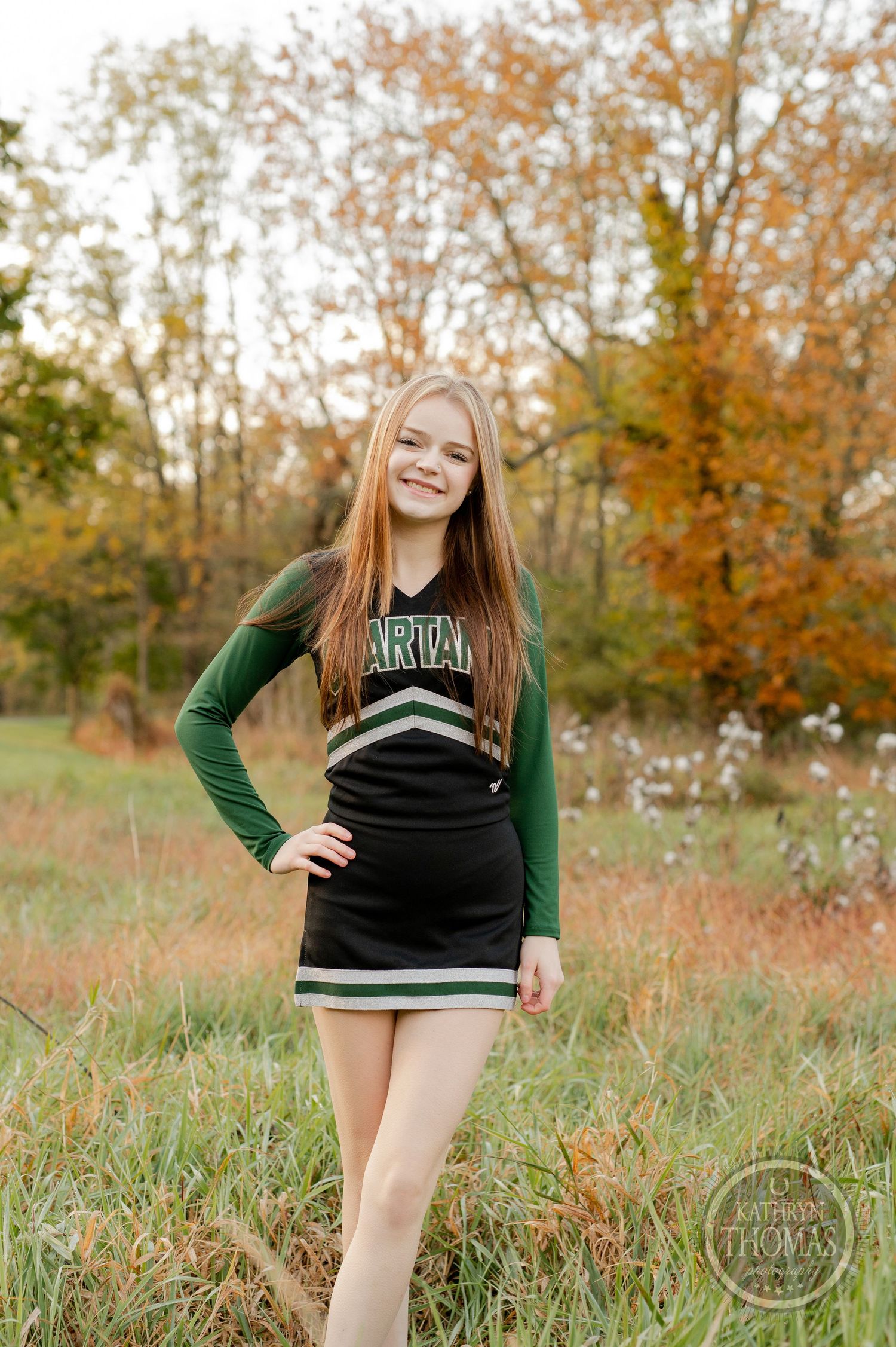 Cheerleader in green and black uniform poses in field with autumn foliage in background.