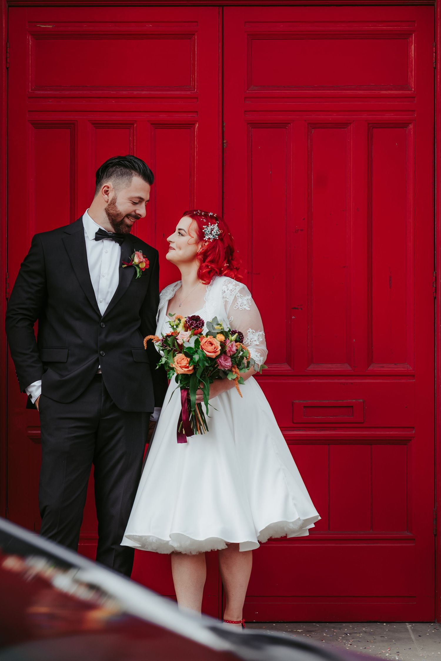 Vintage groom and red head bride in 1950s style dress and black tuxedo in front of bright red door in Bakewell. Burgundy and orange bouquet
