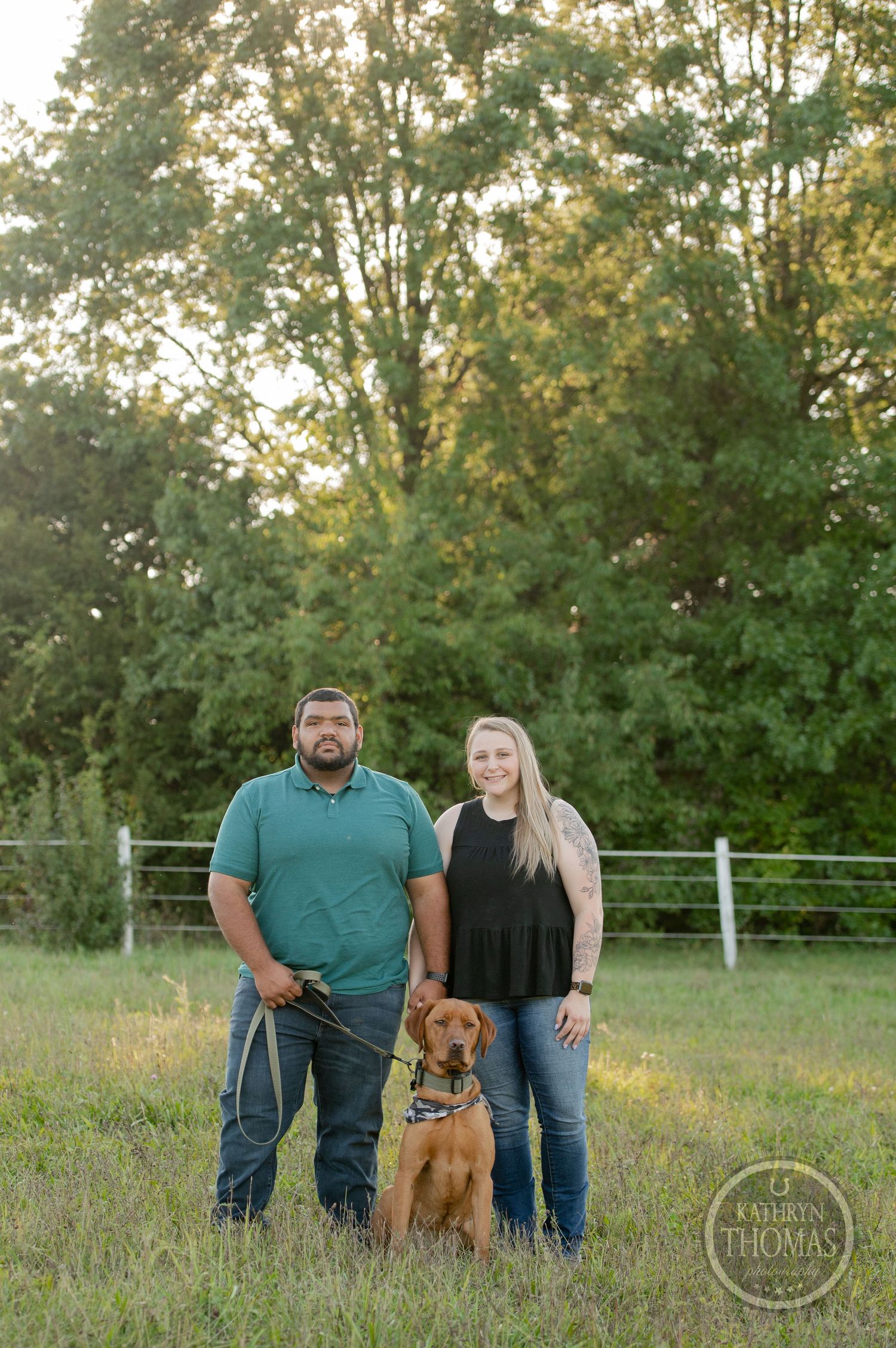 A couple stands with their brown dog in front of a white fence and trees.