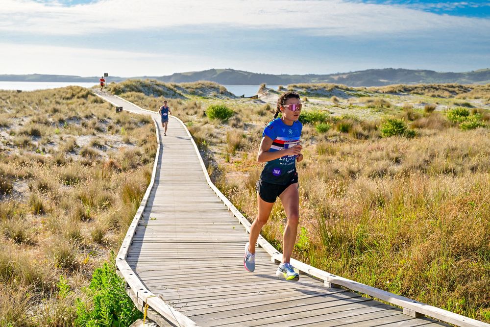 Woman in blue shirt running on wooden boardwalk through coastal sand dunes on a sunny day.