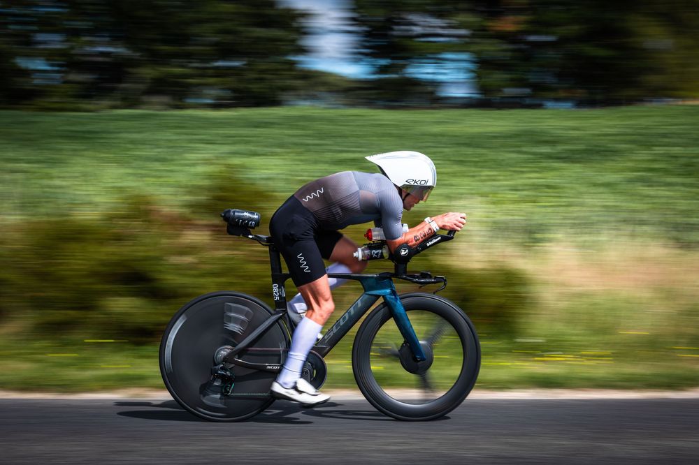 Professional cyclist in aerodynamic position racing on a time trial bike along a scenic road during competition.